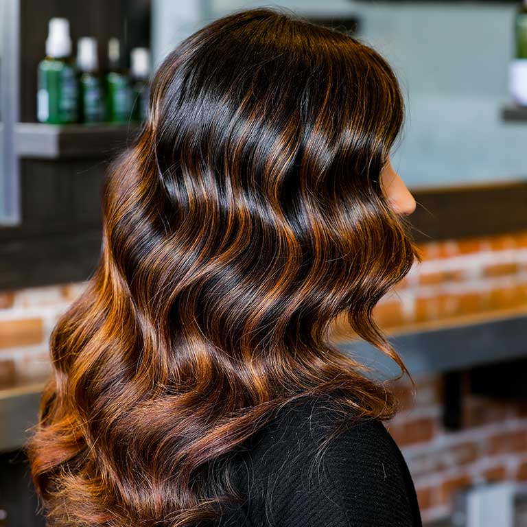 Woman with sleek, dark, curled hair and copper-colored highlights from Cutting Crew Hair Salon.